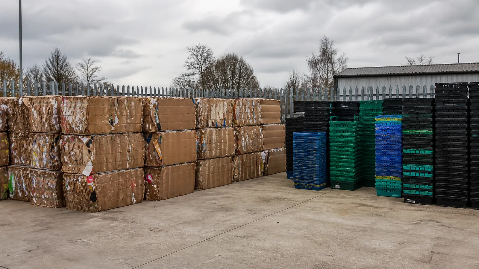 Baled cardboard and stacked empty plastic crates inside a fenced storage compound at a UK recycling yard